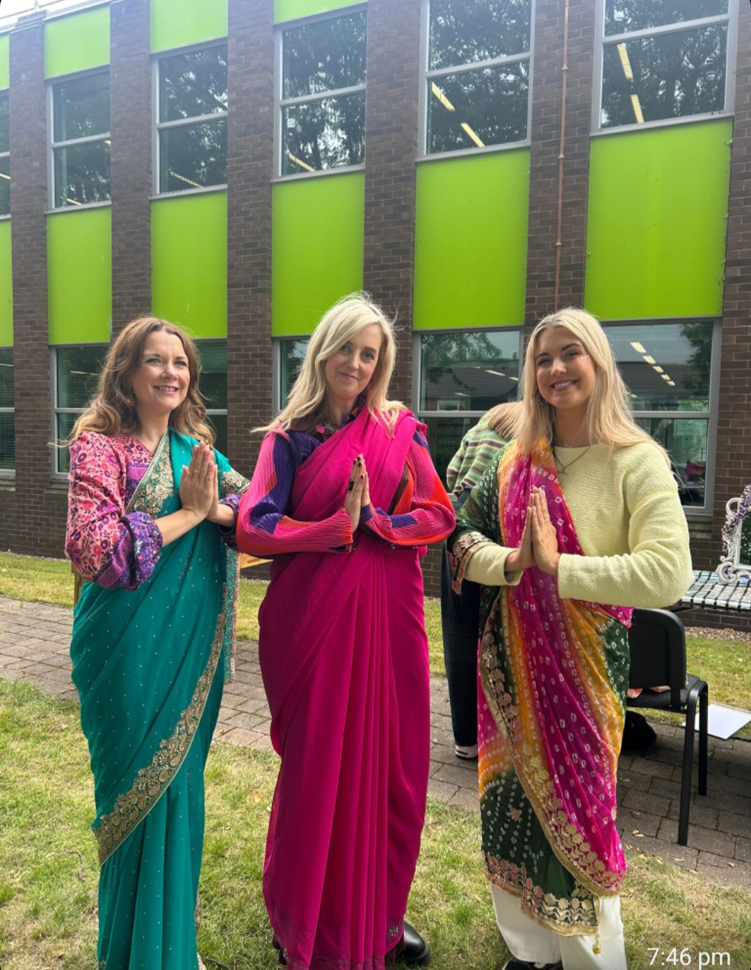 Three adults wearing colourful sarees and traditional South Asian clothing, standing with hands together in greeting outside a school building.